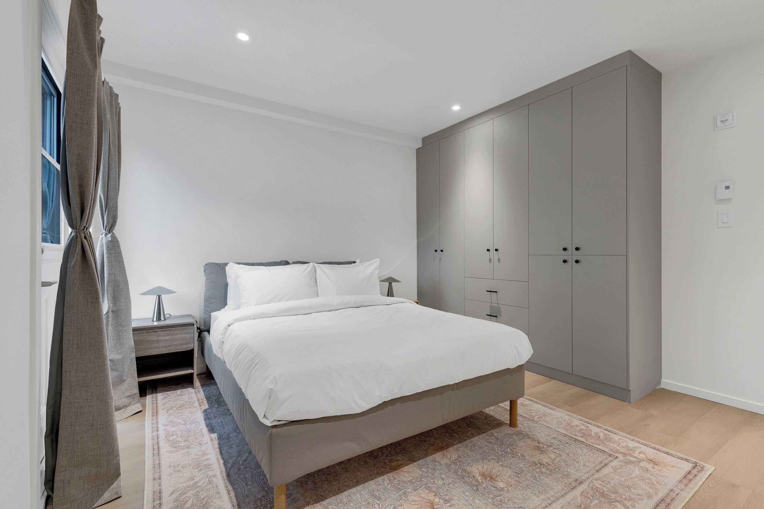 Un dormitorio moderno con una cama grande, un armario gris y una mesita de noche con lámpara.