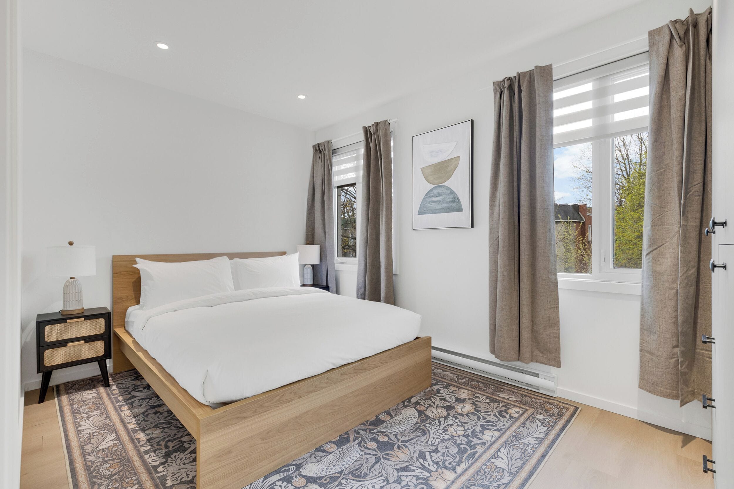 A bedroom with a wooden bed, a nightstand, a patterned rug, and a window with curtains.