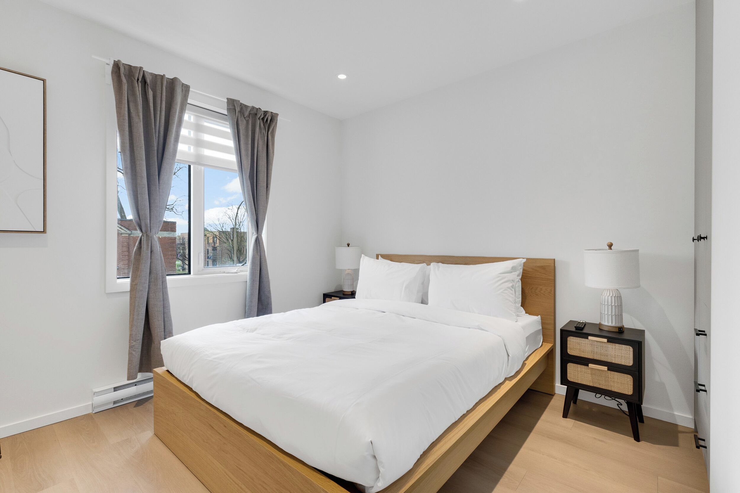 Quarto moderno com cama de madeira, roupa de cama branca, mesa de cabeceira com abajur e janela com cortinas.