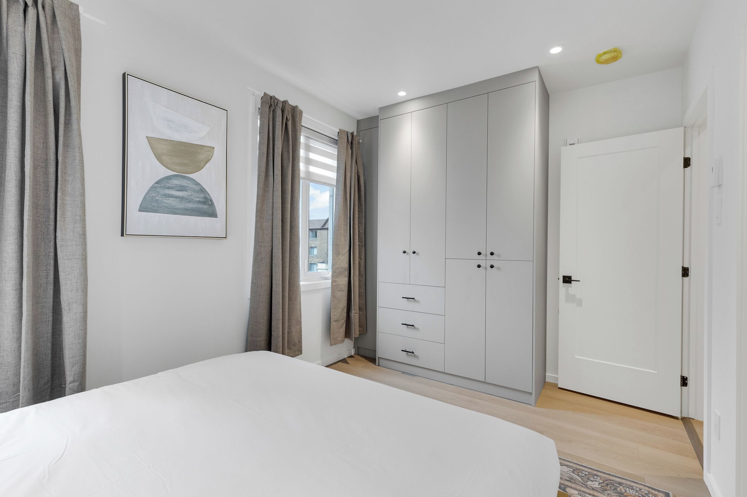 Une chambre à coucher avec un lit, une armoire, une fenêtre avec des rideaux et un tableau encadré au mur.