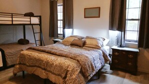 Une chambre avec un lit superposé, un grand lit recouvert d’une couverture à motifs, une table de chevet avec une lampe et une fenêtre avec des rideaux.