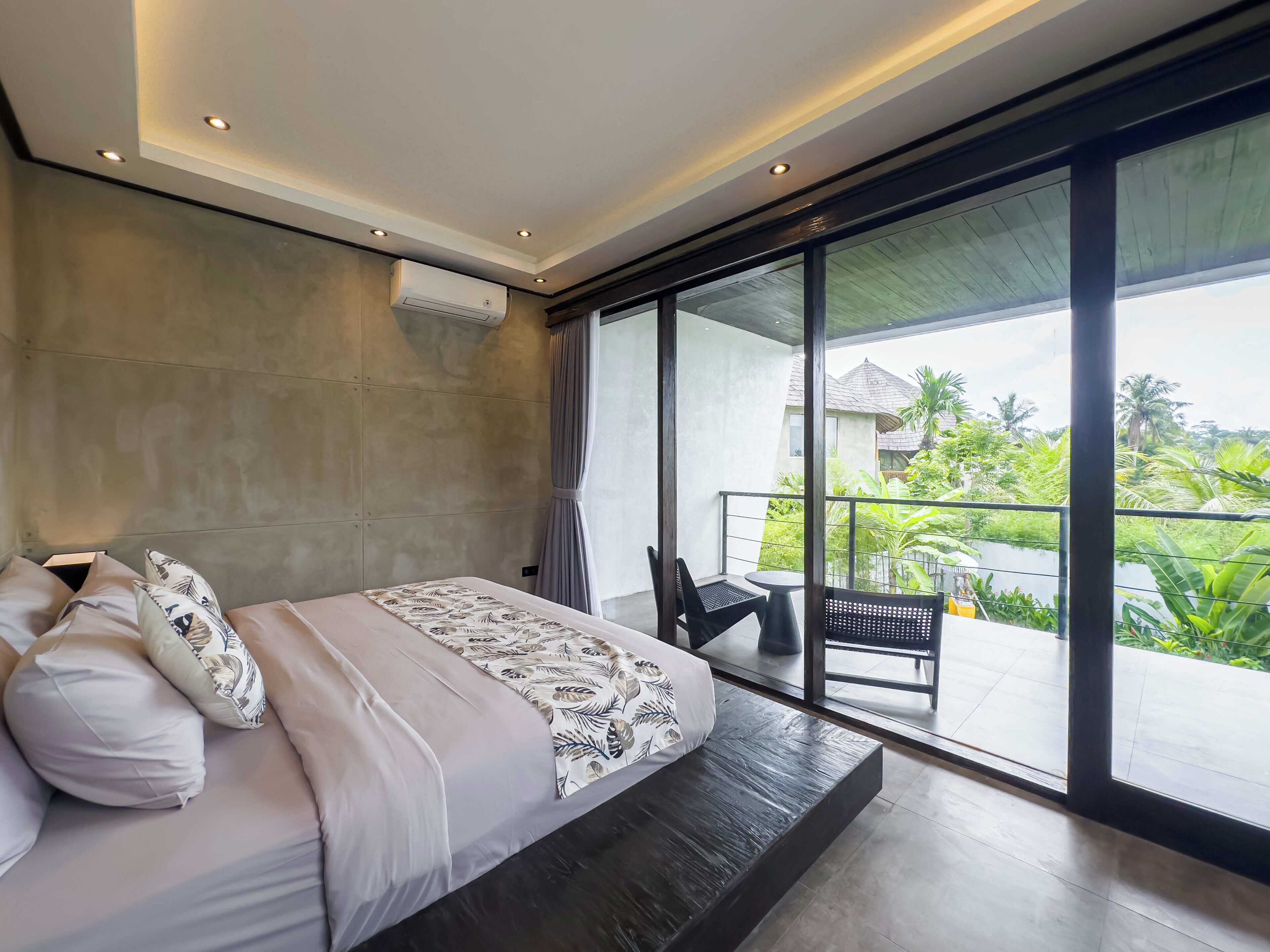 A modern bedroom with a large bed, a balcony with a table and chairs, and a view of greenery.