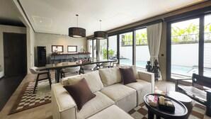 A modern living room with a sofa, coffee table, and dining area. There is a view of a pool and greenery outside. - Iri Villas Ubud  (Kemenuh)