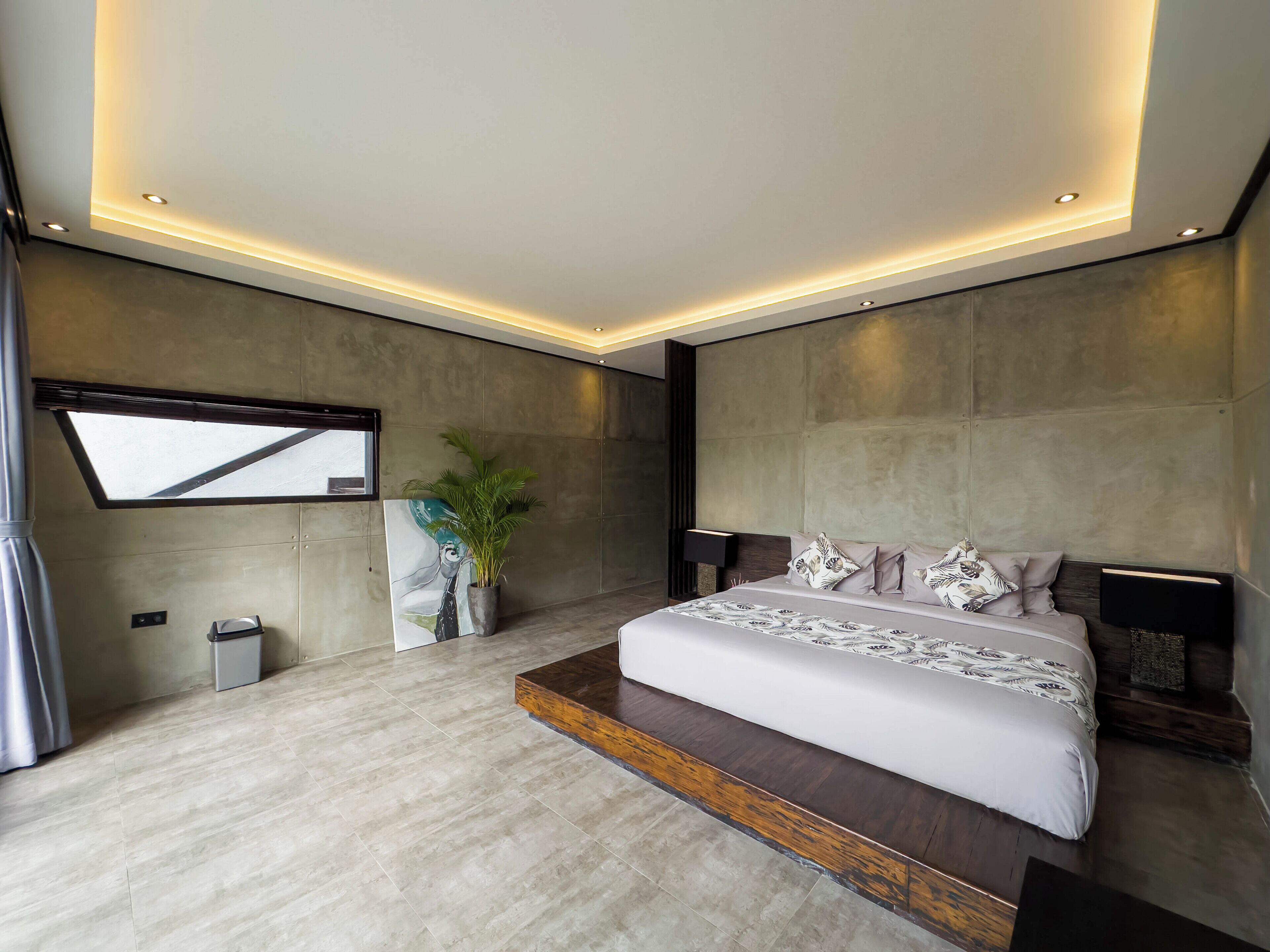 A modern bedroom with a large bed, recessed ceiling lighting, a potted plant, and a large window.