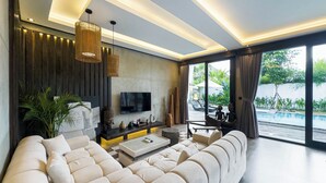 A modern living room with a large beige sofa, a flat-screen TV mounted on the wall, and a large window offering a view of a pool and greenery. - Iri Villas Ubud  (Kemenuh)