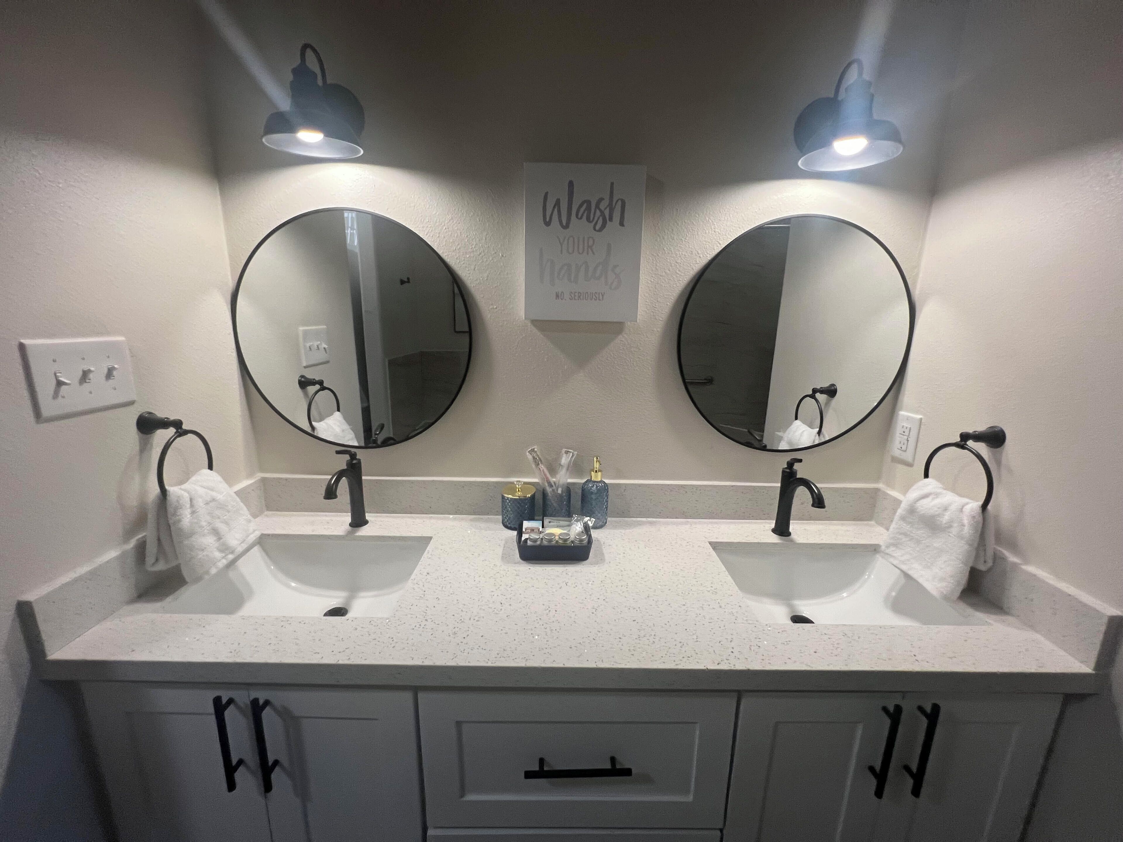 Een badkamer met twee wastafels, twee ronde spiegels en een bordje met de tekst 'Handen wassen. Serieus nu.'