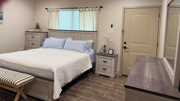 Een slaapkamer met een bed, nachtkastjes, een bankje, een raam met gordijnen, een deur en een plafondventilator.