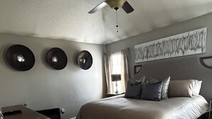 Quarto com cama, escrivaninha, ventilador de teto e uma grande pintura abstrata na parede.