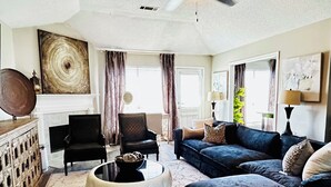 Uma sala de estar espaçosa com ventilador de teto, lareira, um grande quadro abstrato e um conjunto de sofás moderno.