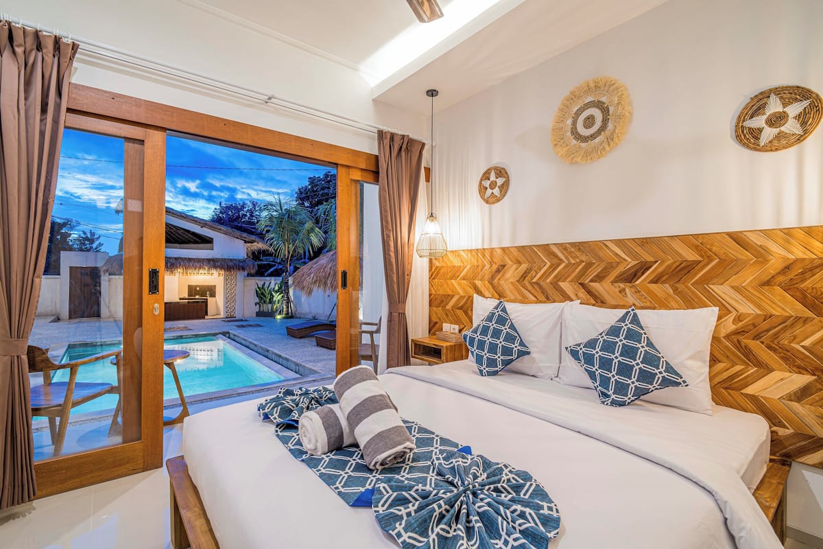 A bedroom with a wooden herringbone headboard, a large bed with patterned bedding, two wall-mounted decorative plates, and a view of an outdoor swimming pool and thatched-roof building.