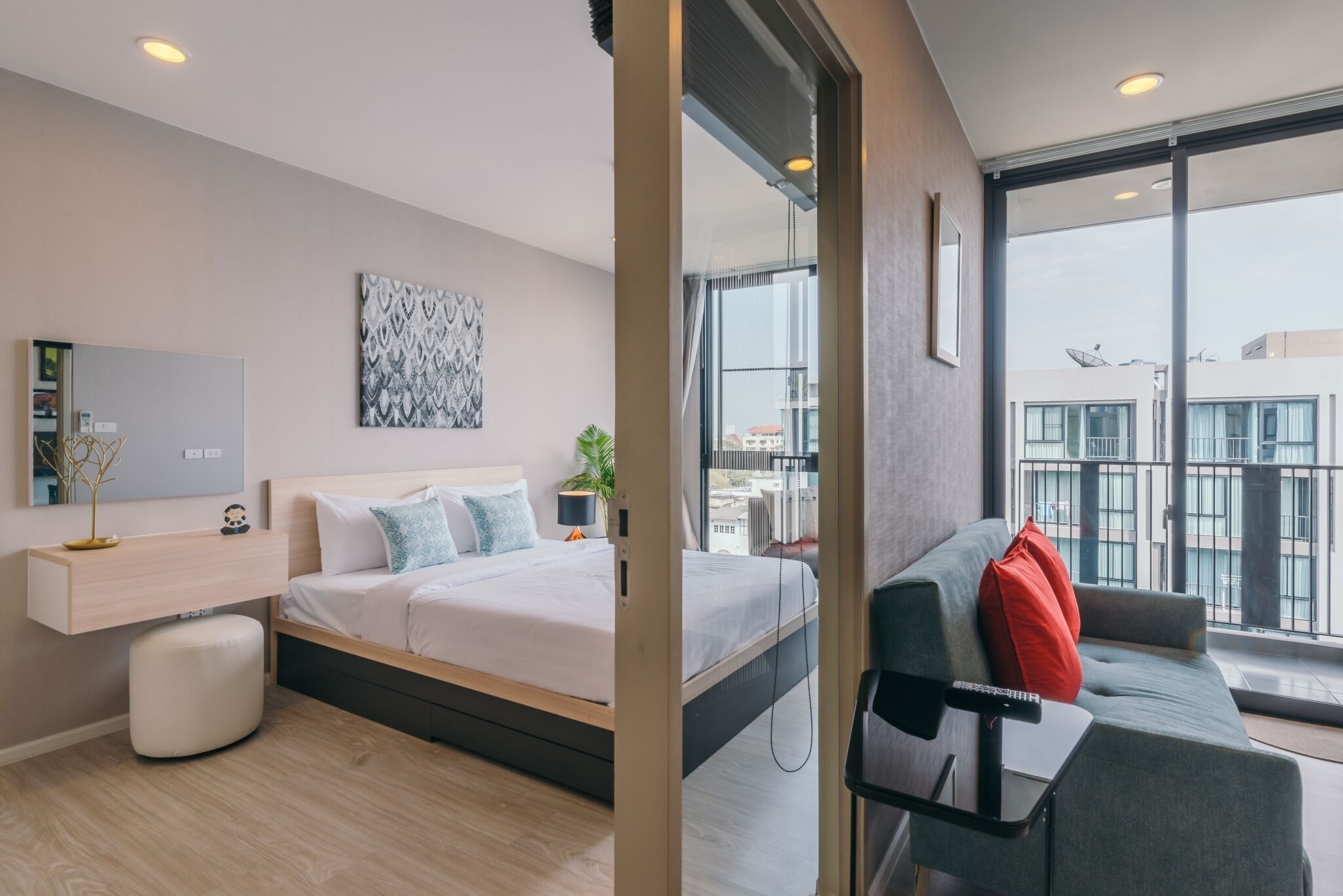 A modern hotel room with a bed, a sofa, a small table, a TV, and a balcony with a view of buildings.