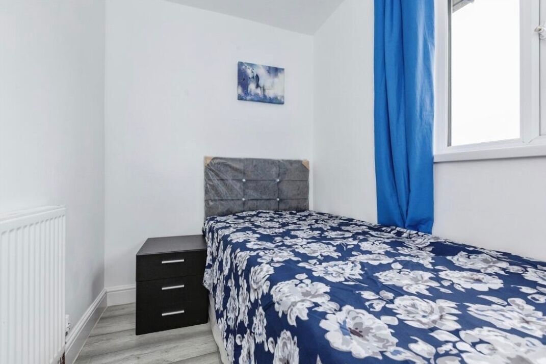 A bedroom with a bed, a nightstand, a dresser, a window with blue curtains, and a wall-mounted picture.