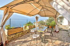 An outdoor patio with a table and chairs, a large umbrella, and a view of the sea and mountains.