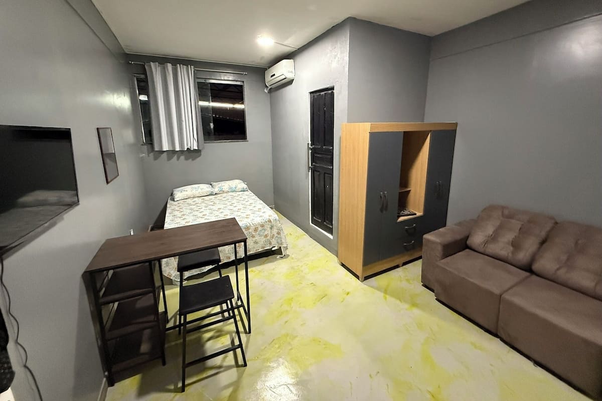 Un espacio habitable compacto con una cama, un sofá, un escritorio con silla, un armario y un televisor montado en la pared.