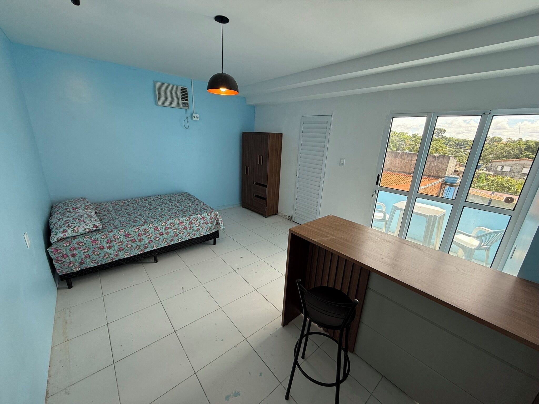 Una habitación con cama, escritorio, silla, armario y ventana con vista a edificios.