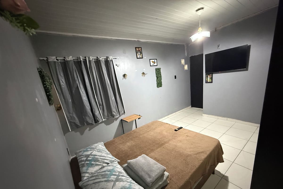 Un dormitorio con una cama, una mesita, un televisor montado en la pared y una cortina.