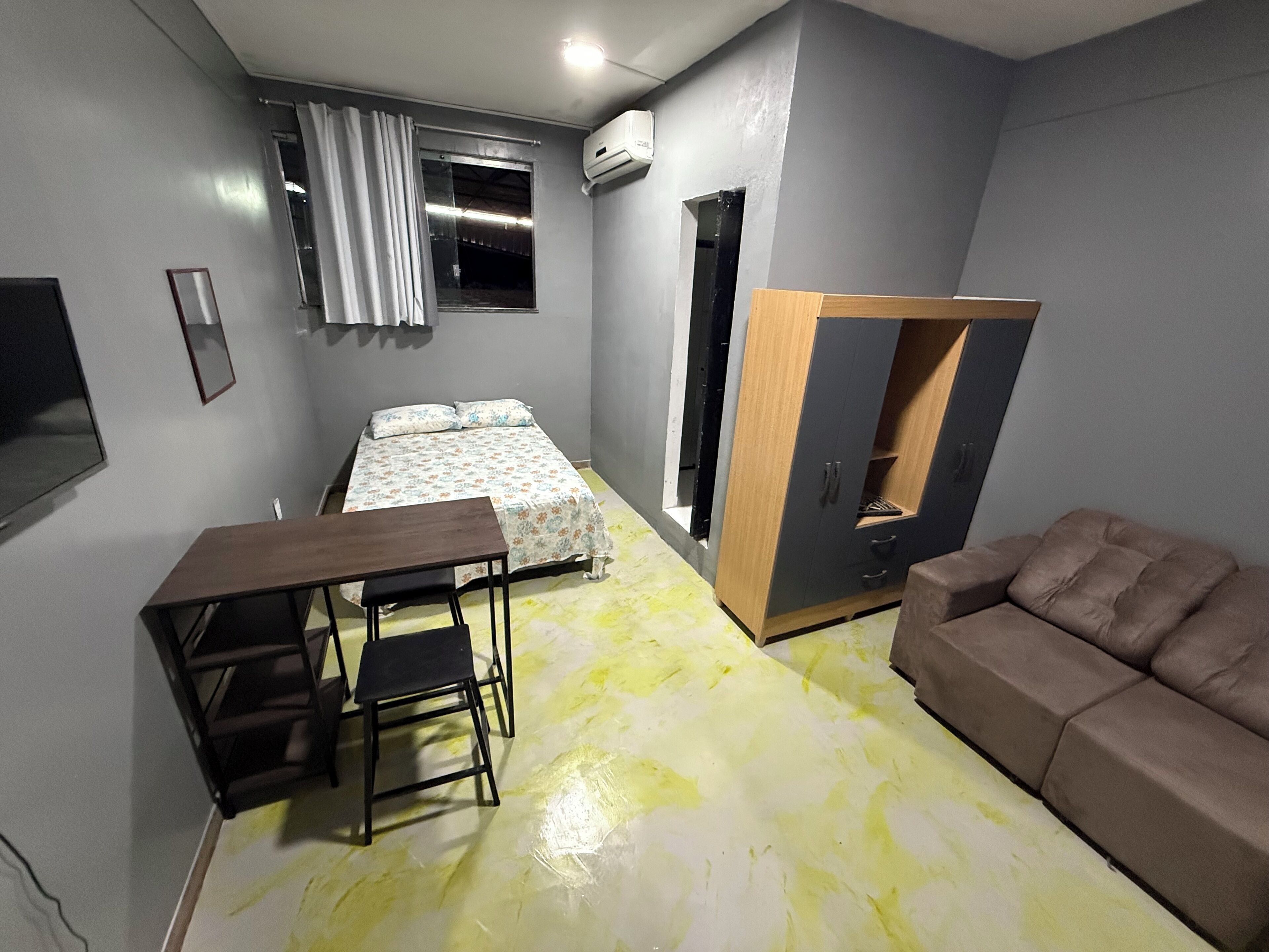 A compact bedroom with a bed, a wooden wardrobe, a brown sofa, and a small table with a chair.