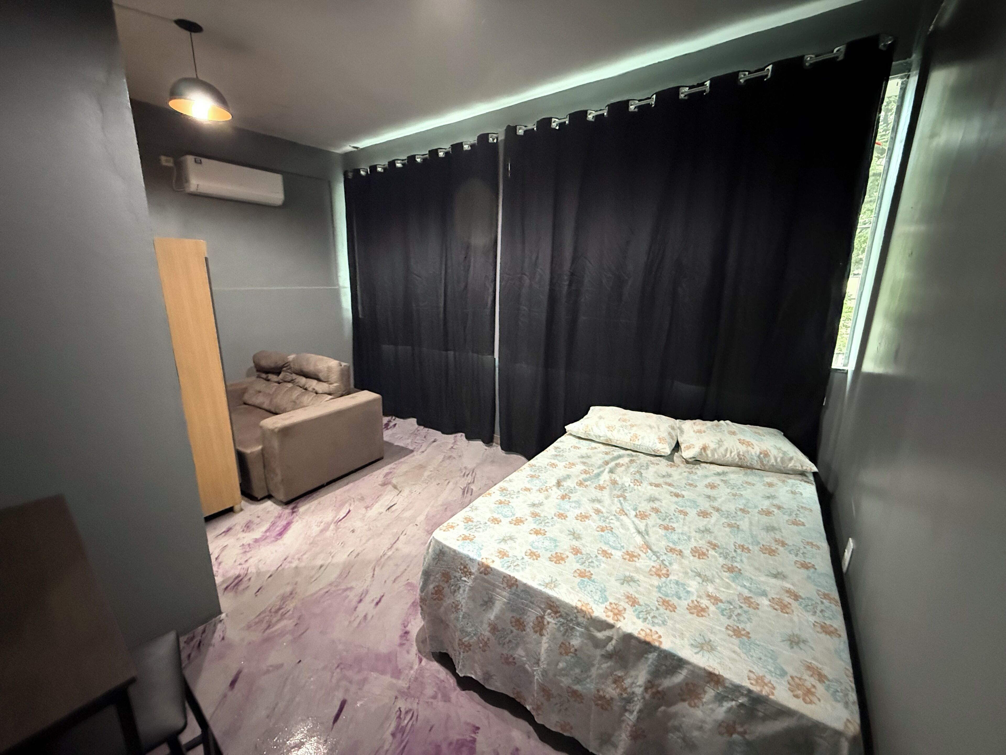 A bedroom with a bed, a sofa, a window with curtains, and an air conditioning unit.