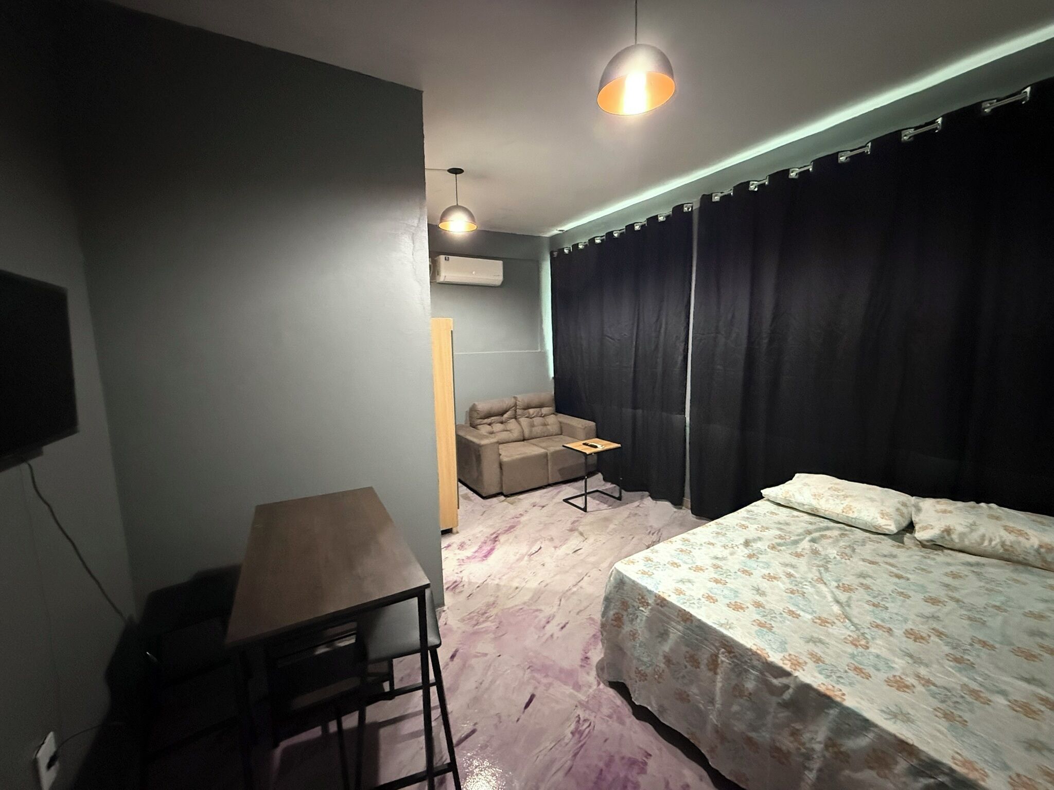 A bedroom with a bed, a desk, and a chair. There is a window with curtains and an air conditioning unit on the wall.