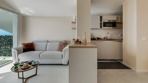 Una sala de estar moderna con un sofá, una mesita de centro con tazas de té y pasteles, y un área de cocina con microondas y armarios.
