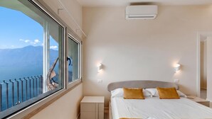 Una habitación de hotel con una cama, una mesita de noche, un ventanal con vistas a montañas y un equipo de aire acondicionado.