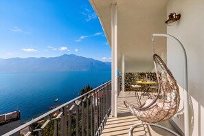 A balcony with a view of the sea and mountains, a hanging chair, and a small table with a chair.
