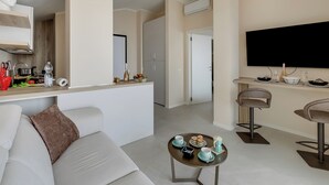 Una sala de estar moderna con un sofá, una mesa de centro con tazas de té, una zona de cocina con mostrador y un televisor montado en la pared.