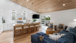 A modern kitchen with wooden cabinets, a dining area with chairs, a blue sofa, and a large window overlooking a deck.