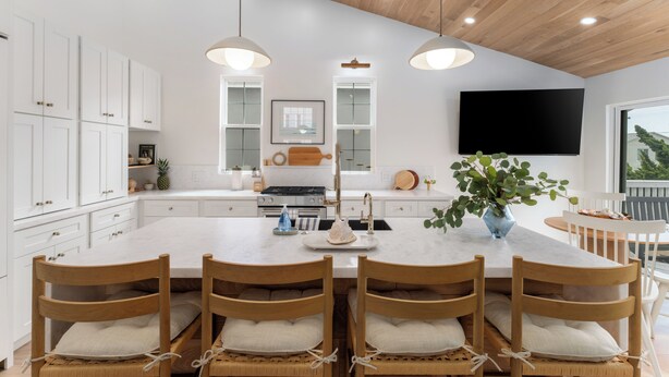 A modern kitchen with white cabinets, a large island with seating, pendant lighting, and a flat-screen TV mounted on the wall.