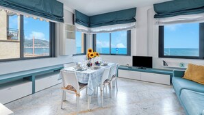 Een moderne eetruimte met een tafel voor vier personen, grote ramen met uitzicht op de oceaan en een blauwe bank met een geel kussen.