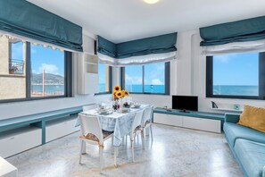 A modern dining area with a table set for four, large windows with ocean views, and a blue sofa with a yellow pillow. - Front Beach Magic Sea - Sea Front, Laigueglia, Italy (Laigueglia)
