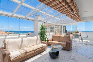 Un balcon spacieux doté d’un plafond en verre, d’un canapé beige, d’une table basse en verre et d’une plante en pot.