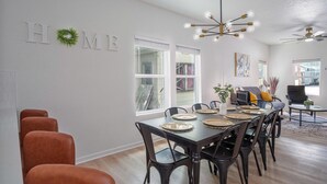 Un espace repas avec une longue table dressée pour six personnes, entourée de chaises, et agrémenté d’un ventilateur de plafond et de luminaires suspendus.