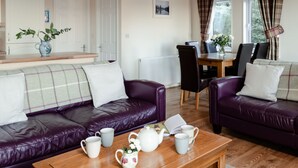 A cozy living room with a purple leather sofa, a wooden coffee table with tea set, and a dining area with a table and chairs. - Lodge 2 by Interhome (Boat of Garten)