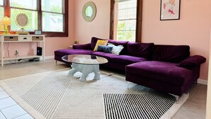 Un salon avec un canapé violet, une table basse en verre et un tapis blanc. Il y a deux fenêtres avec des stores, une petite table d’appoint et des œuvres d’art accrochées au mur.