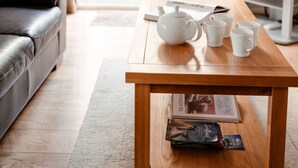 A cozy living room with a wooden table, a white teapot, and three white cups. A black leather sofa is against the wall, and a television is on a stand to the right. - Lodge 7 by Interhome (Boat of Garten)