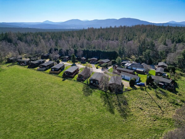An aerial view of a rural housing development with multiple wooden houses, surrounded by green fields and forested hills.