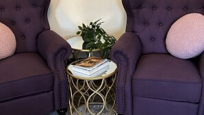 Deux fauteuils violets à dossier capitonné, une petite table ronde avec un pied doré, une plante et une pile de livres.