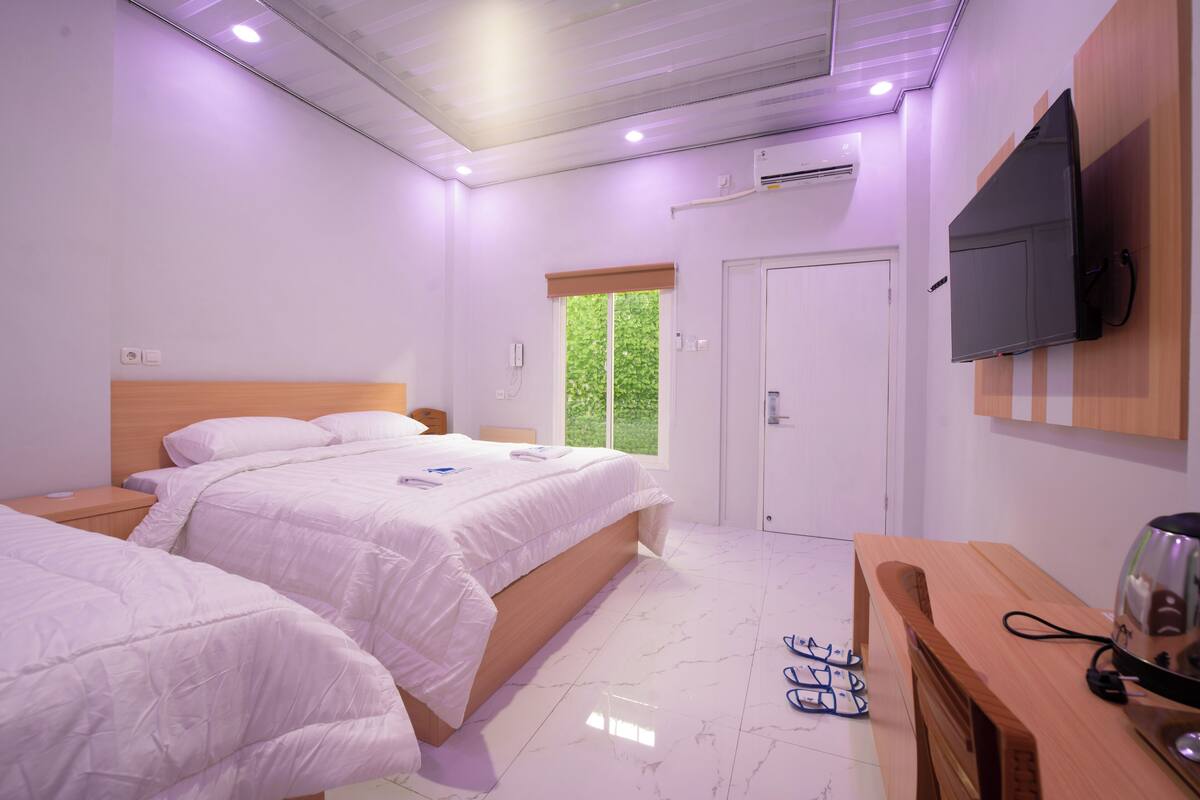 Een slaapkamer met twee bedden, een houten bureau, een televisie en een raam met uitzicht op groen.