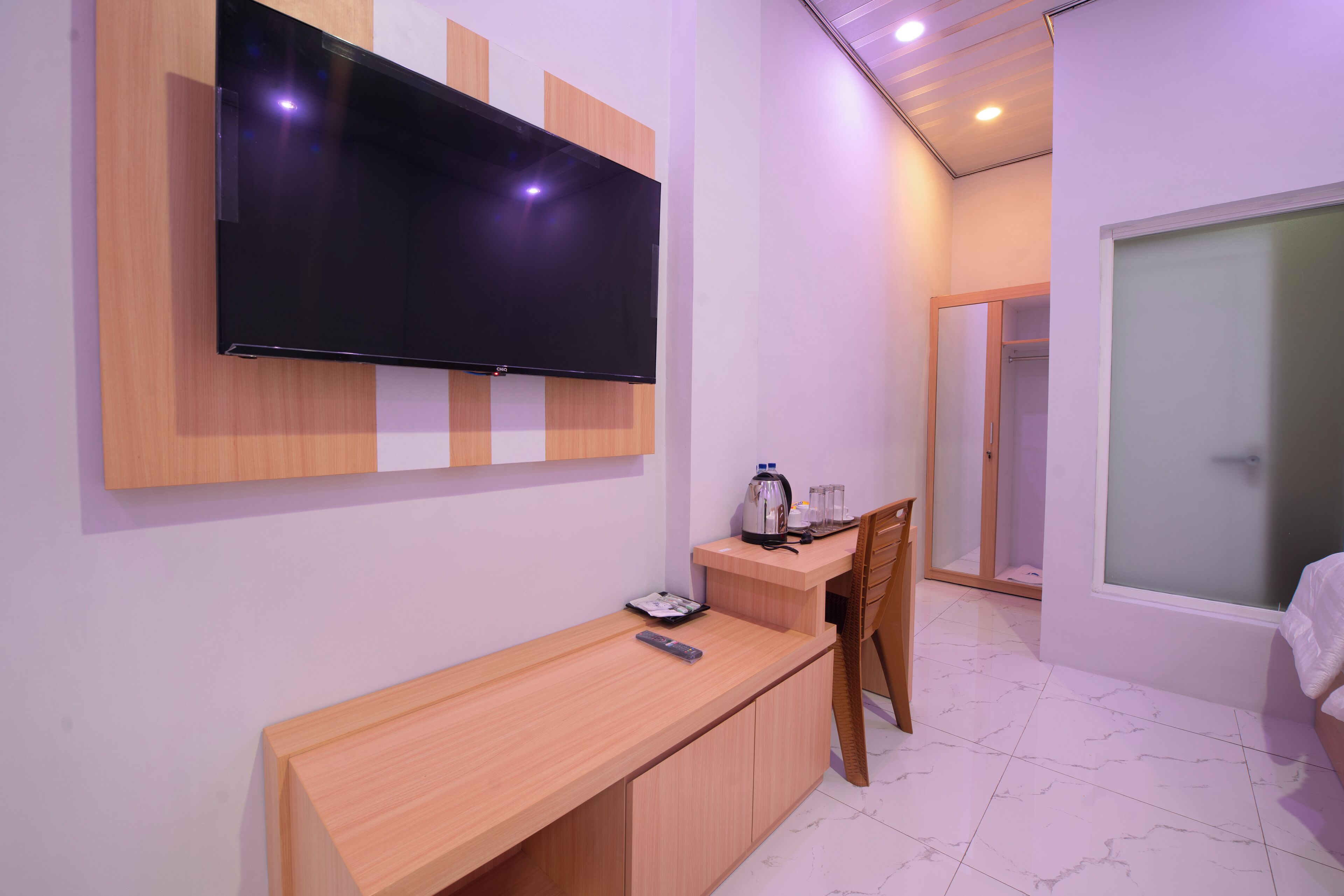 Un dormitorio moderno con una televisión de pantalla plana montada en la pared, un escritorio de madera con un control remoto, una cafetera y una silla.