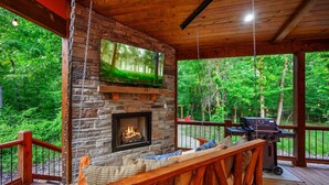 A covered deck with a stone fireplace, a television, and a grill.