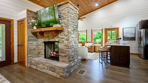 A stone fireplace with a TV above it, a kitchen area with a refrigerator, and a seating area with a view of greenery outside.