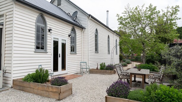 Una casa blanca con un patio de grava, terraza de madera y macizos de flores con diversas plantas.