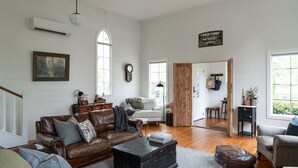 Un salón amplio con un sofá de cuero, un sillón blanco y una mesa de centro de madera. Un reloj y un cuadro decoran las paredes.
