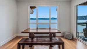 Un coin repas avec une table et un banc en bois, une fenêtre donnant sur un lac et des montagnes, et un balcon avec des sièges d’extérieur.