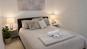 Una cama bien hecha con una colcha acolchada y varias almohadas, dos lámparas de noche, una pequeña planta en maceta y una obra de arte en la pared sobre la cama.