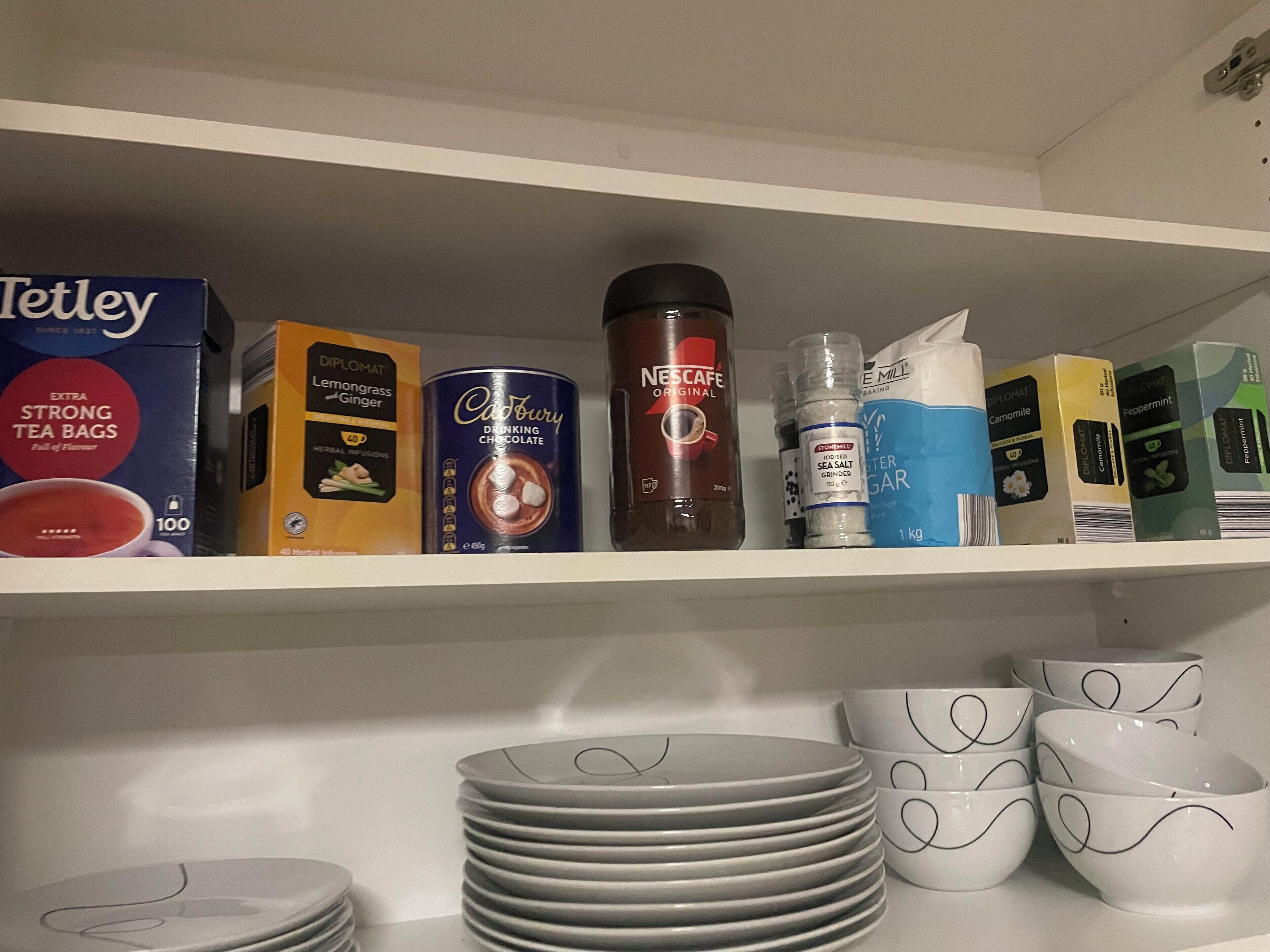 Uma estante com vários saquinhos de chá, uma lata de chocolate quente Cadbury, uma cafeteira Nescafe e alguns ingredientes de confeitaria. Abaixo da estante, pratos e tigelas empilhados.