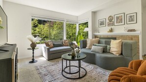 Ein modernes Wohnzimmer mit einem grauen Sofa, einem runden Couchtisch und einem großen Fenster mit Blick auf Grünflächen.