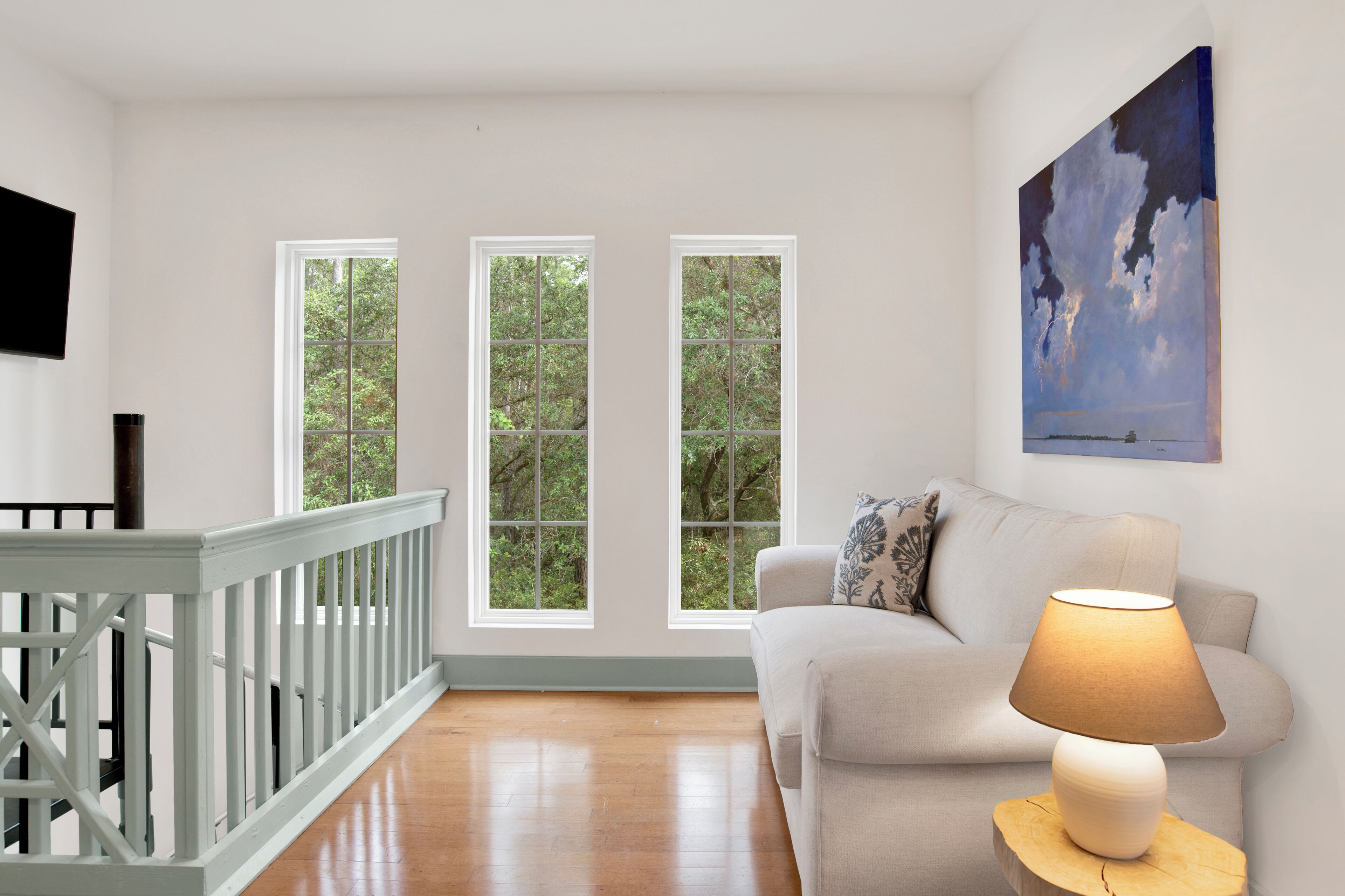 A room with a wooden floor, a white sofa, a lamp on a wooden side table, and three windows with a view of greenery.