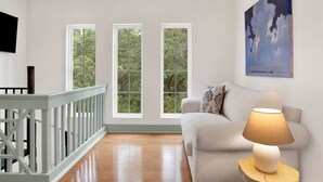 A room with a wooden floor, a white sofa, a lamp on a wooden side table, and three windows with a view of greenery. - "Idlehour" new cottage in Fairhope, AL - NEW LISTING (Fairhope)